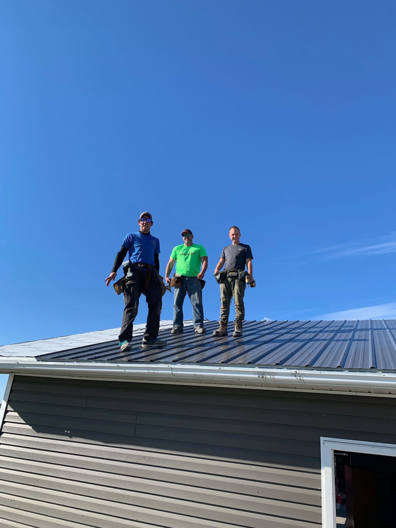 A three-man crew standing on a finished dark metal roof under a clear blue sky